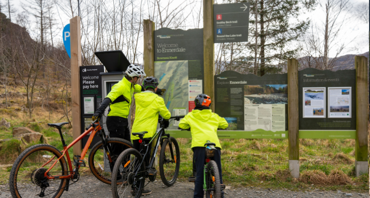 A dad and two boys with bikes stood looking at information board in car park