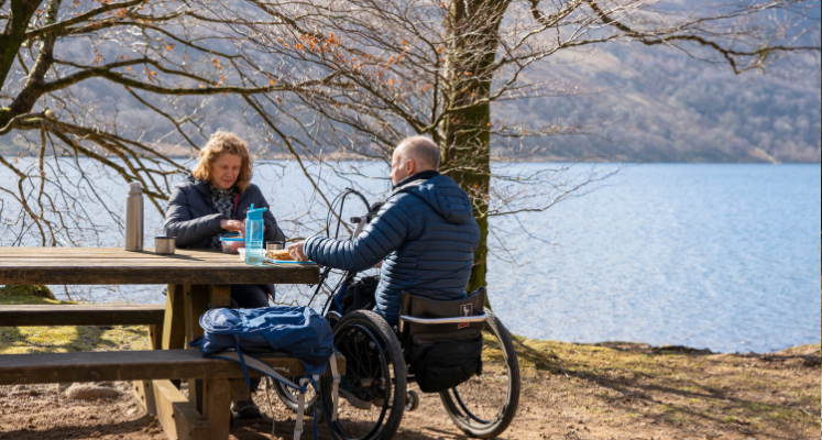 Man in wheelchair with wife sat at picnic bench by the lake shore having lunch