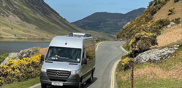 Shuttle bus on a narrow road with a lake and steep mountains behind