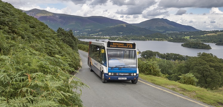 Stagecoach bus on the road next to a lake with fells in the background.