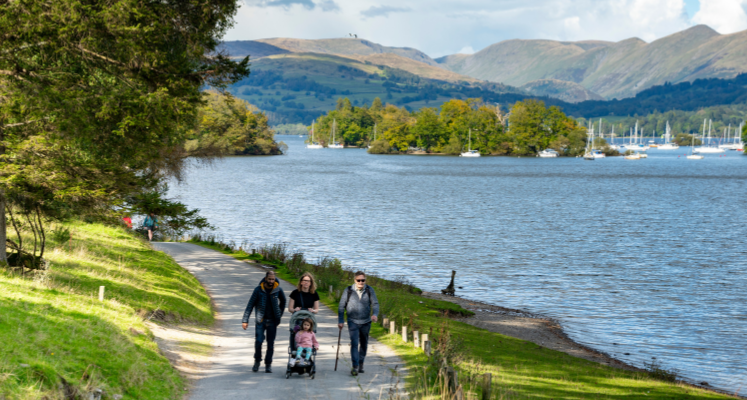 A family pushing a pram with grandad with walking stick walking along path by Windermere's shore