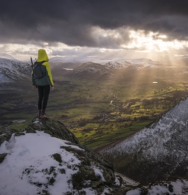 A walker standing on the top of a fell looking out onto a snowy Lake District.