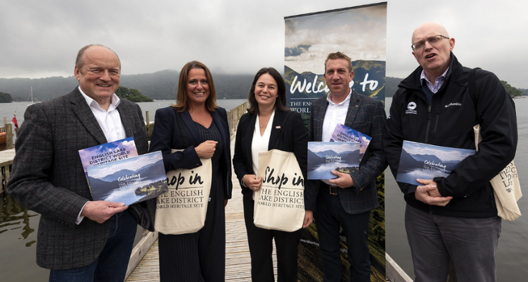 Tree men and two women stood on a jetty holding celebrating the Lakes items with a banner and a lake behind them