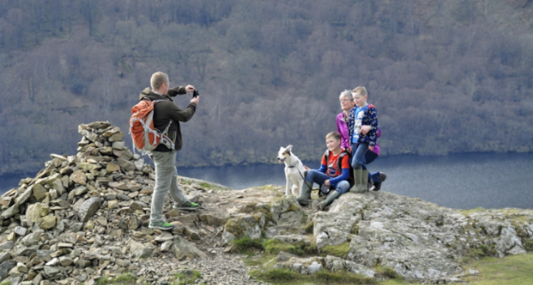 A family taking a photo on the top of a fell there is a man taking the photo with a woman, dog and two children posing for a photo with a lake a trees in the background