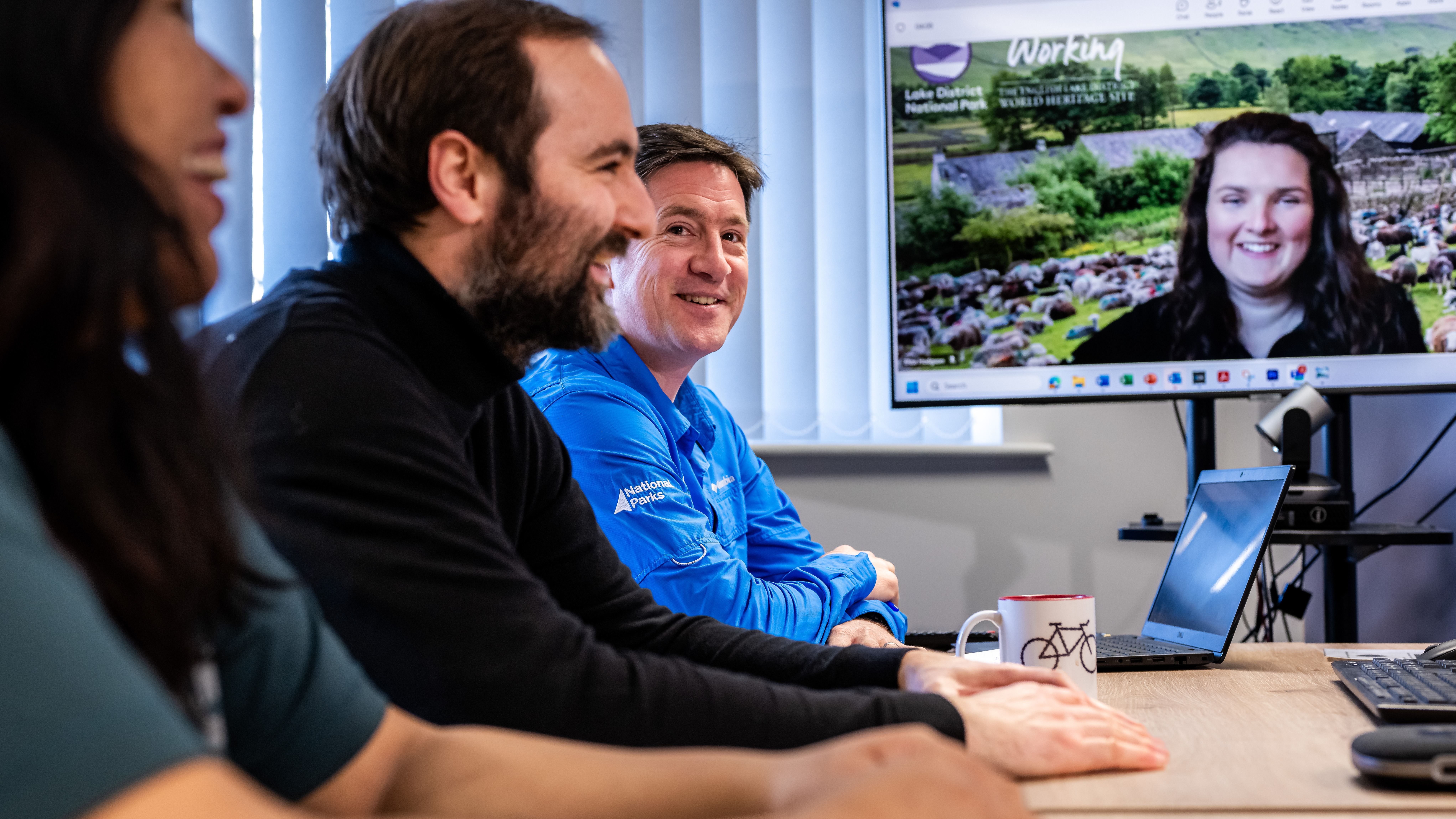 Three staff members in a meeting room. a large screen shows the forth staff member who is working from home and joins the meeting virtually.
