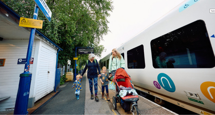 Grandma, mum and two toddlers and a baby in a pram walking by a train smiling