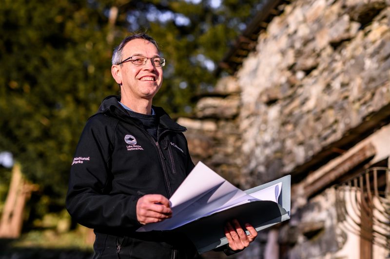 Male Planner standing outside a traditional lakeland building. He is wearing a black uniform jacket which show the LDNPA and National parks logos.