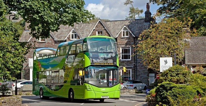 The 599 open top bus driving round a corner. There is a grey slate house in the background, and green trees and bushes on either side of the road.