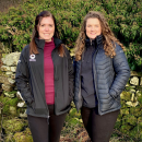 Farming officers Claire on the left and Eliza on the right stood smiling in front of a wall and hedge in their Lake District National Park uniform