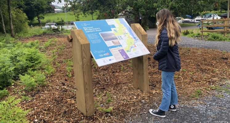 A lady stood in a black coat, jeans and black trainers reading a sign in the Lake District by a car park