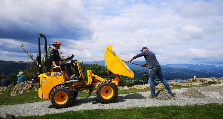 Two Field Rangers on Orrest Head one in a yellow dumper and other one raking out gravel onto the path with lakeland mountains in background