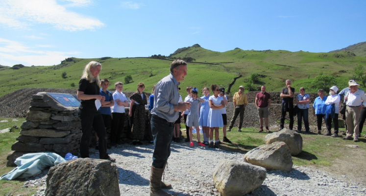 A group standing round listening to a Lake District talk amongst green hills and blue skies
