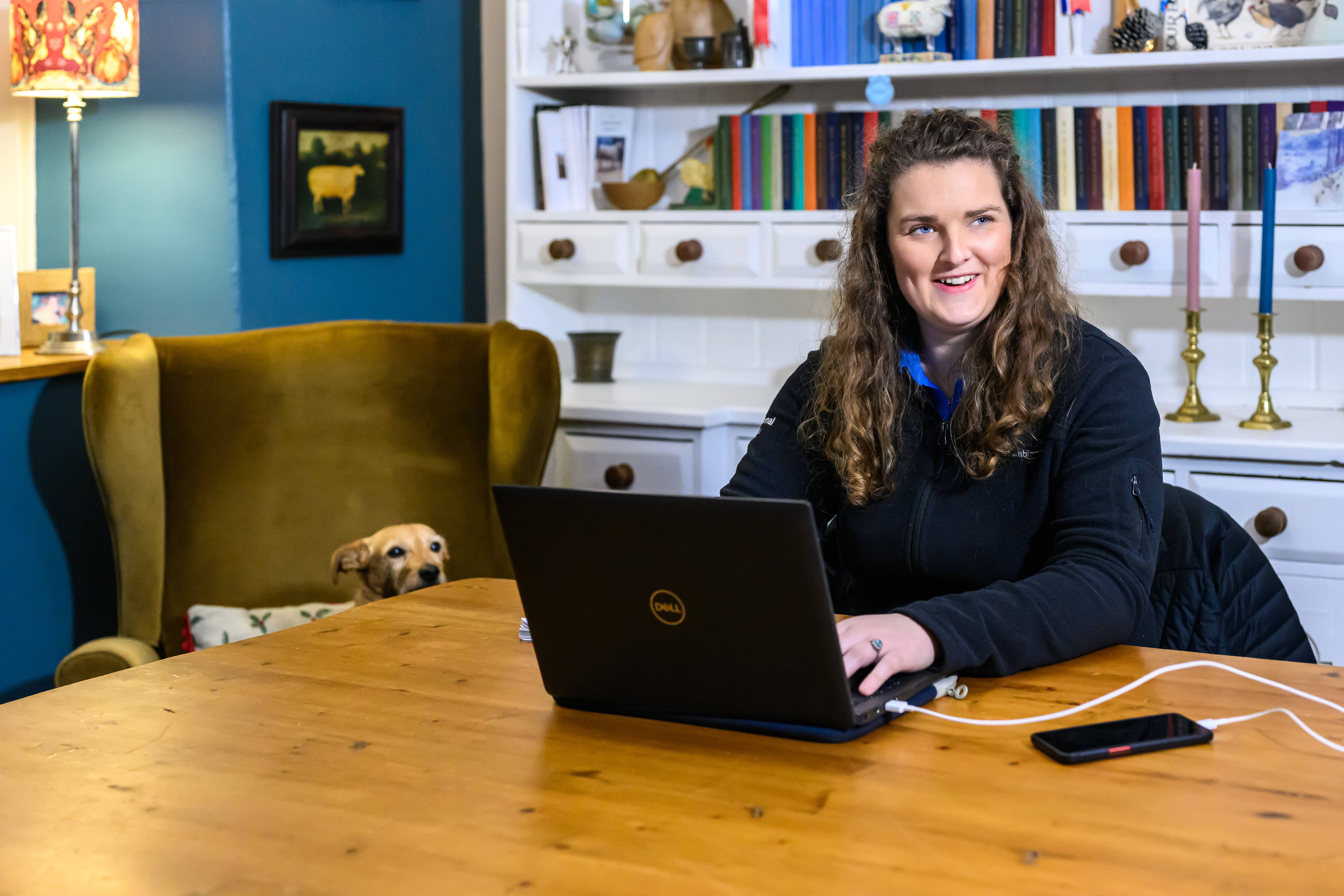 National park staff member wroking form home at her kitchen table. a dog's head can be seen peeping out from an armchair. A large white dresser with books, is in the background  