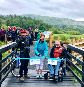 The official opening of teh West Windermere Way with a man on a bike, a lady and a man in a wheelchair smile at the camera holding a blue ribbon. There is a group of people looking on behind them 